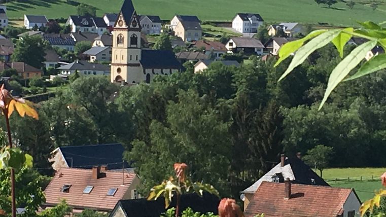 Blick auf ein Dorf mit Kirche und Häusern, umgeben von grünen Hügeln und Bäumen.