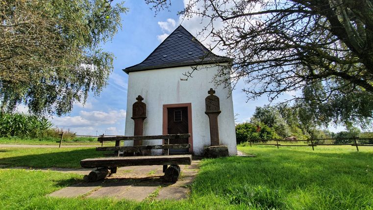 Une petite chapelle blanche avec un toit en ardoise, entourée d'arbres et d'un banc au premier plan.