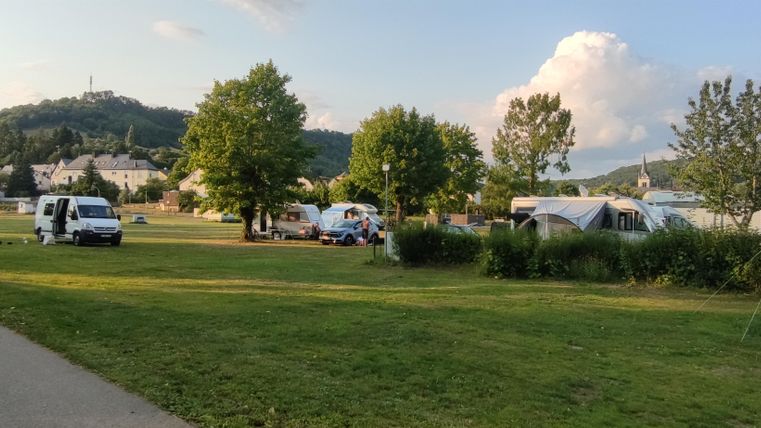Un terrain de camping avec des zones herbeuses et de nombreux arbres. À l'arrière-plan, on peut voir des camping-cars et des tentes.