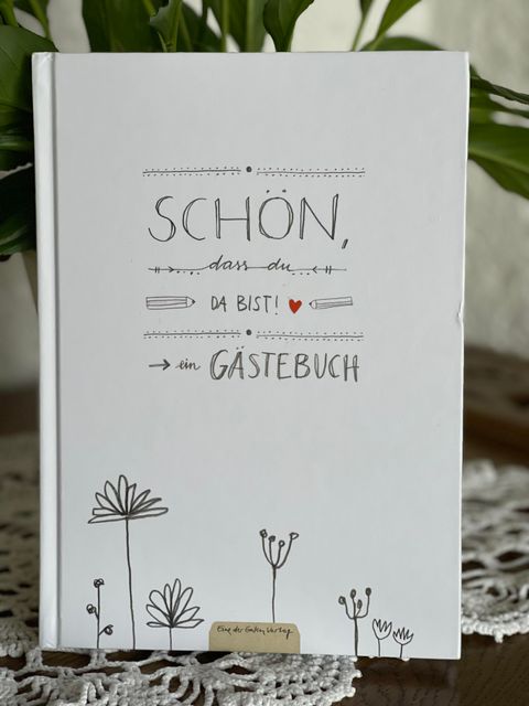 Ein einladendes Gästebuch mit der Aufschrift "SCHÖN, dass du da bist!" und einer einfachen Zeichnung von Blumen. Es liegt auf einem Tisch, umgeben von einer dekorativen Häkelarbeit.