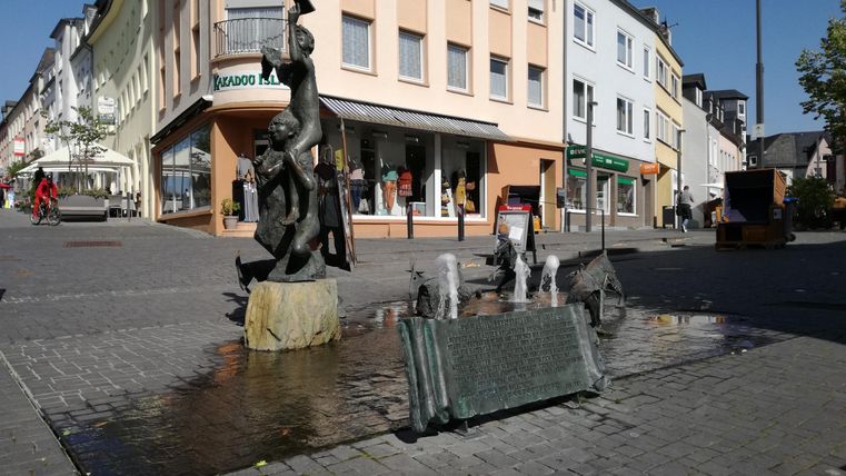 Eine belebte Straße mit einem Brunnen und einer auffälligen Statue. Im Hintergrund sind verschiedene Gebäude zu sehen.