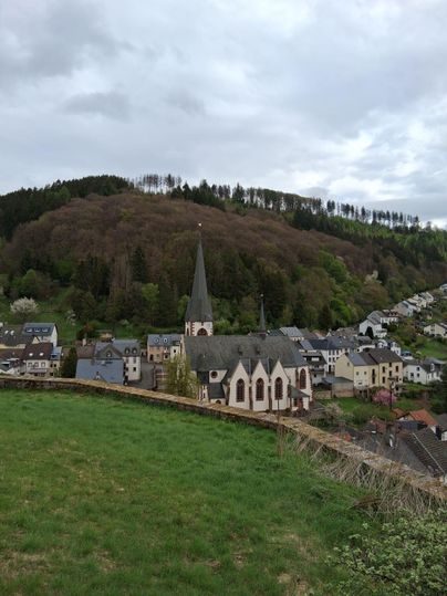 Ein malerisches Dorf mit einer Kirche und umgeben von bewaldeten Hügeln. Der Himmel ist bewölkt und die Landschaft wirkt ruhig und einladend.