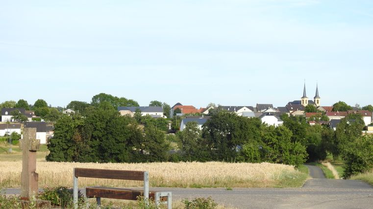 Landschaft mit Bank, Feld und Dorf im Hintergrund.