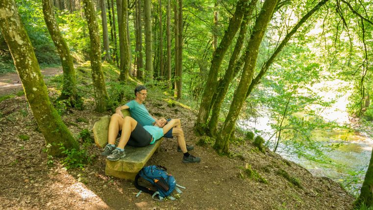 Ein Paar sitzt auf einer Bank im Wald, umgeben von Bäumen und einem Fluss im Hintergrund.