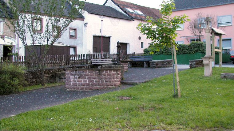 Dorfplatz in Gindorf mit Brunnen, Bänken und Häusern im Hintergrund.