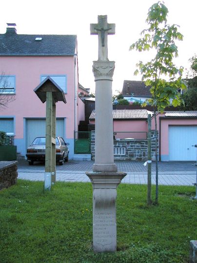 Wegekreuz auf einem Dorfplatz mit einem Baum und einem Haus im Hintergrund.