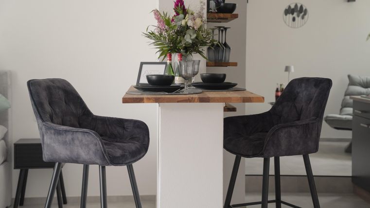 Ein moderner Essbereich mit zwei bequemen, grauen Hockern und einem rustikalen Tisch. Auf dem Tisch stehen Teller und eine Vase mit frischen Blumen.