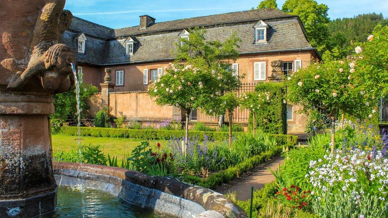 Ein schöner Garten mit bunten Blumen und einem Brunnen im Vordergrund. Im Hintergrund steht ein charmantes Haus mit grünen Bäumen.