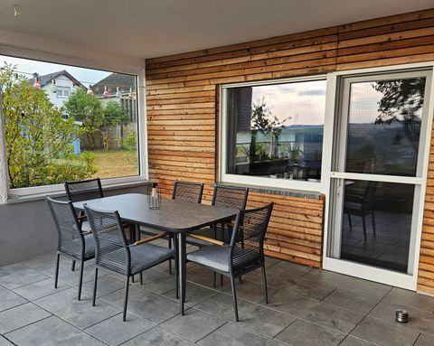 A cozy outdoor area with a table and chairs on a terrace. One can see large windows and a green surroundings.