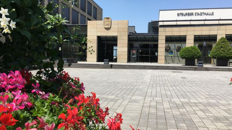 Ein modernes Gebäude mit großen Fenstern steht im Vordergrund. Im Vordergrund blühen bunte Blumen und es ist ein klarer blauer Himmel zu sehen.