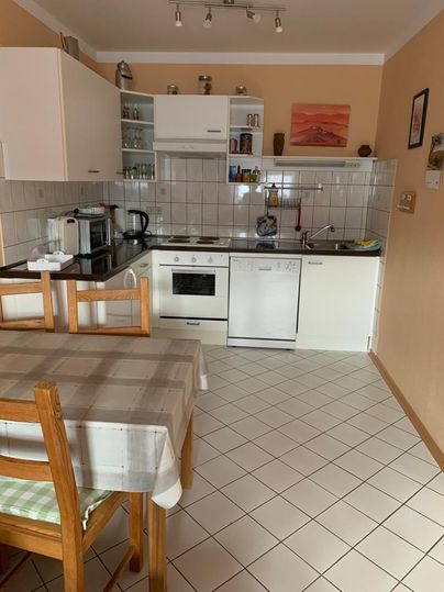 Eine helle Küche mit modernen Geräten und einer Esstischgruppe. Die Wände sind in einem warmen Farbton gehalten, und die Fliesen sind neutral.