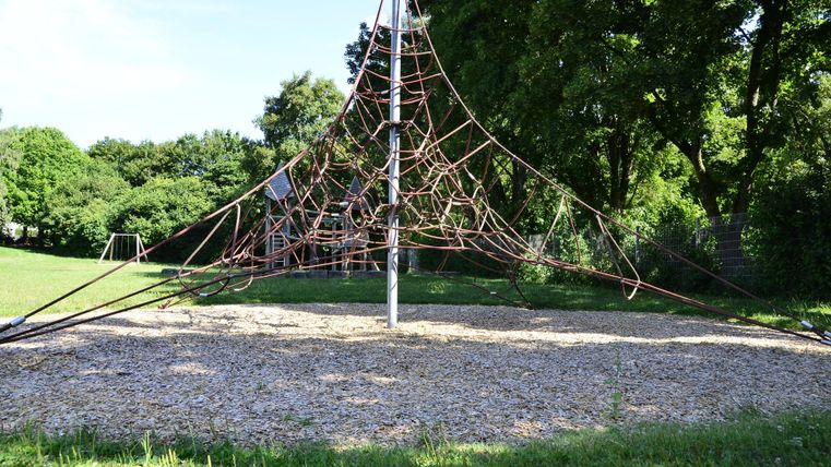 Un terrain de jeux avec une structure d'escalade en cordes se trouve sur une pelouse. À l'arrière-plan, des arbres et de l'herbe verte sont visibles.