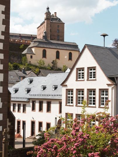 Schloss Malberg im Hintergrund mit modernen Gebäuden und blühenden Büschen im Vordergrund.