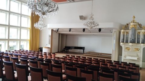 A large event room with many chairs and a stage area. The walls are light and there are elegant chandeliers.