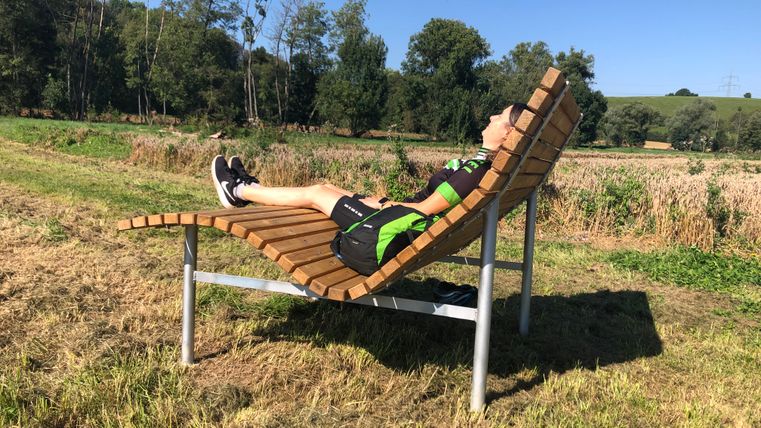 Person liegt auf einer geschwungenen Holzbank in einer ländlichen Umgebung.