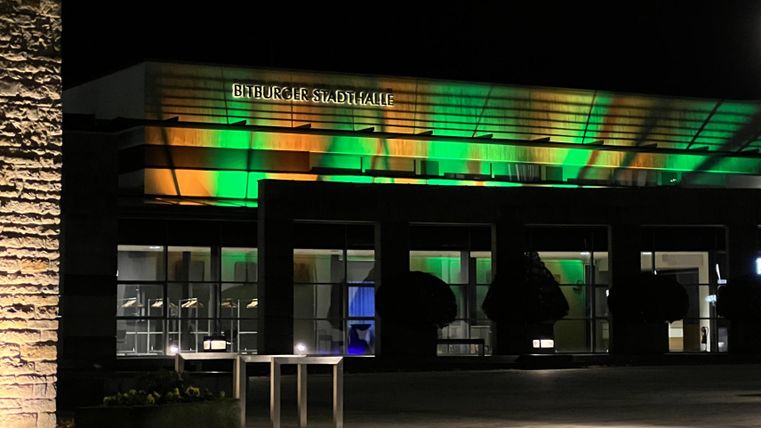 Ein modernes Gebäude bei Nacht, beleuchtet mit grünen und orangefarbenen Lichtern. Die Fassade ist schlicht und ansprechend gestaltet.