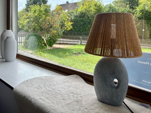 A stylish lamp with a round gray base and a reed-made lampshade is standing on a table. Outside, there is a green meadow and a fence visible.