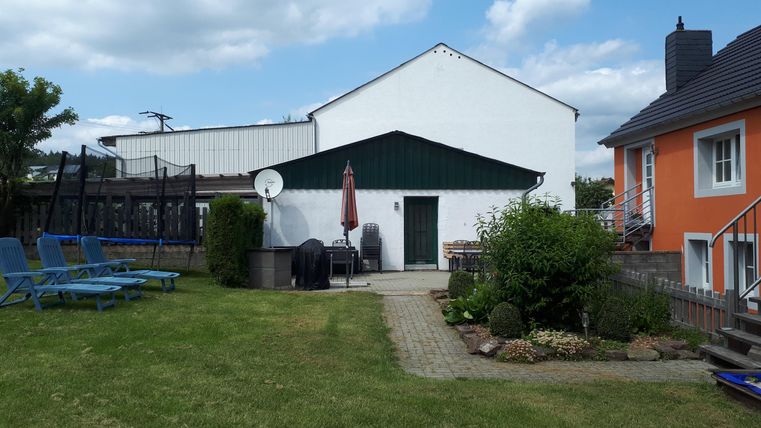 Ein ruhiger Garten mit Rasenfläche, Sonnenliegen und zwei Gebäuden. Der Himmel ist blau mit einigen Wolken.