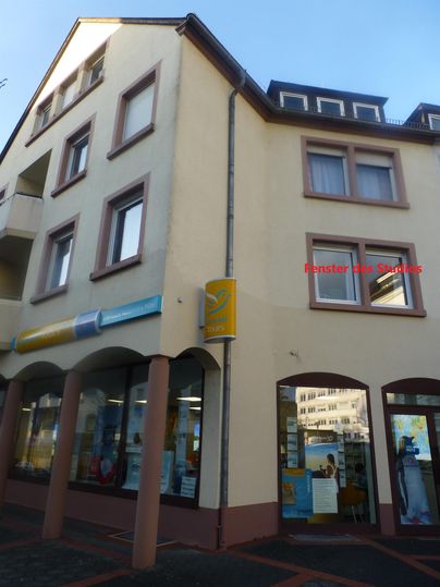 Ein dreigeschossiges Gebäude mit einem gelben Schild. Im Erdgeschoss sind große Fenster und der Eingang sichtbar.