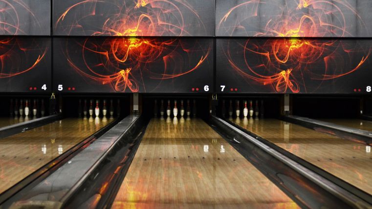 Une piste de bowling avec des surfaces en bois brillantes et des tableaux d'affichage électroniques en arrière-plan. Les murs sont décorés d'un design moderne et abstrait aux couleurs chaudes.
