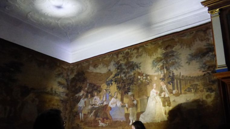 A historical room with a decorative mural. People are standing in the foreground and admiring the artistic depiction.