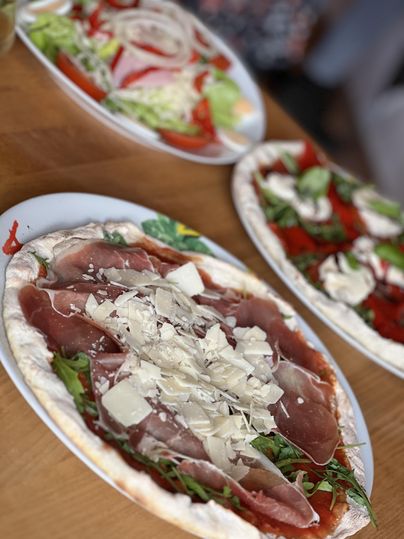 Eine köstliche Pizza mit Schinken und Rucola liegt auf einem Tisch. Im Hintergrund sind frische Salate zu sehen.