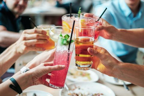 Several hands hold different cocktails as they toast over the table. In the background, food and guests can be seen, creating a cozy atmosphere.