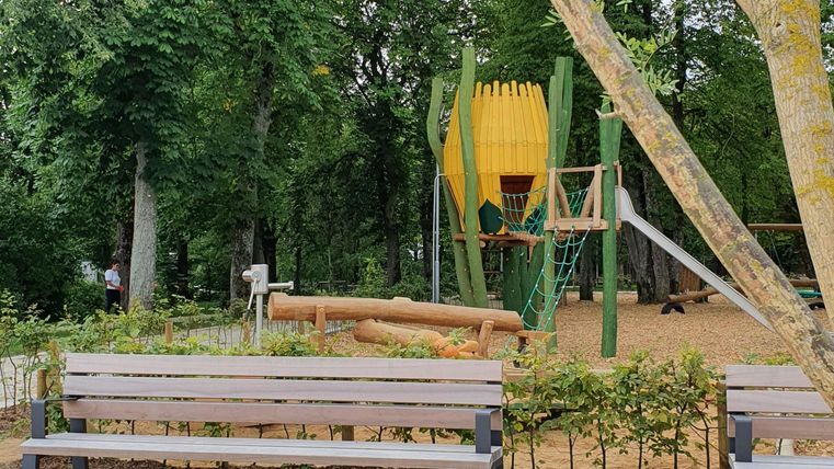 Ein Spielplatz in einem Park mit grünen Bäumen. Im Hintergrund steht eine farbenfrohe Spielkonstruktion und Bänke laden zum Sitzen ein.
