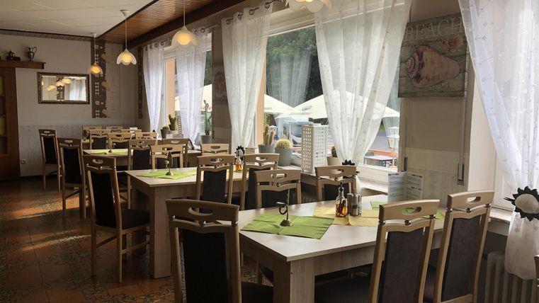 Ein gemütliches Restaurant mit Holzstühlen und Tischen. Große Fenster sorgen für viel Tageslicht und Blick auf die Außenterrasse.