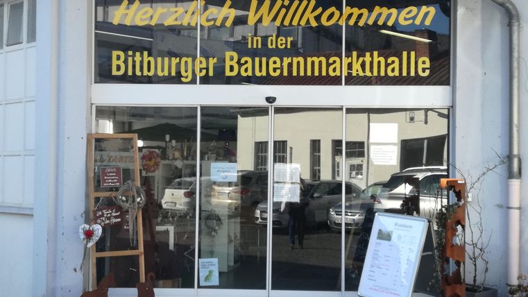 Ein einladender Eingang zur Bitburger Bauernmarkthalle. Die Schaukästen an der Seite zeigen Informationen und Dekorationen.