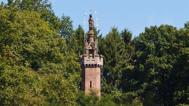 Une haute tour, couronnée d'une statue, s'élève au milieu d'une verdure luxuriante. Le ciel bleu confère à la scène une atmosphère accueillante.