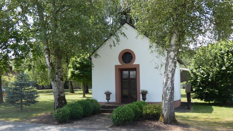 Kleine weiße Kapelle umgeben von Bäumen und Sträuchern.