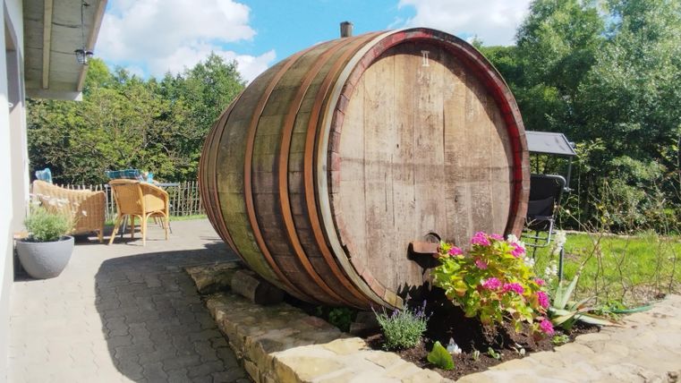 Ein großes Holzfass steht im Garten, umgeben von bunten Blumen. Im Hintergrund sind bequeme Gartenmöbel und Bäume zu sehen.