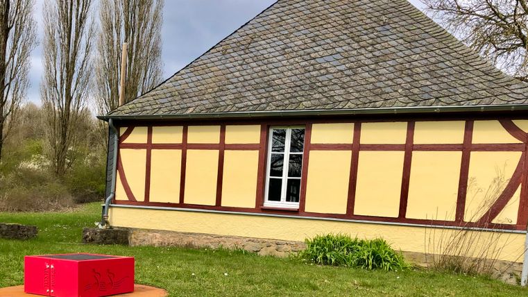 Ein historisches Gebäude mit einem schrägen Dach und bunten Wänden steht im Hintergrund. Im Vordergrund sieht man eine rote Box auf einem orangefarbenen Fußboden.