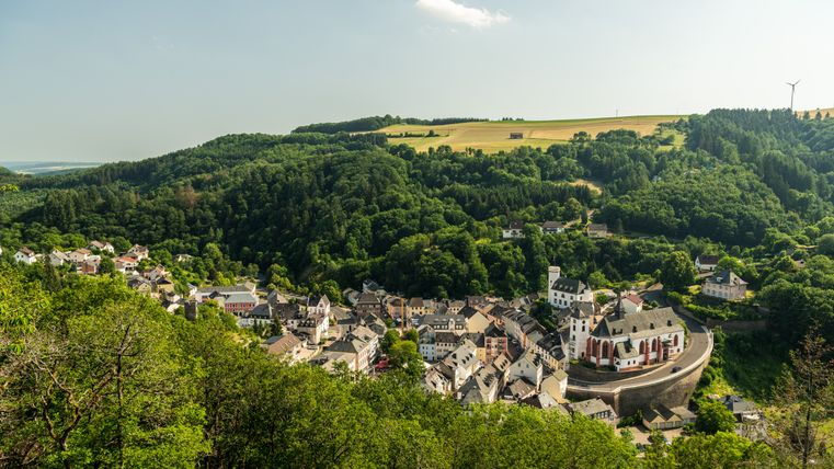 Panoramablick auf Neuerburg, umgeben von grünen Hügeln und Wäldern, mit einer Kirche im Vordergrund.