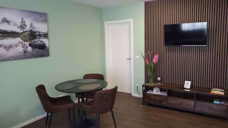 Un salon moderne avec une table ronde, des chaises et une télévision. Un tableau d'un paysage est accroché au mur.