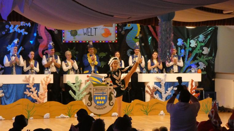 A lively stage scene with a dancer in costume. In the background, several people are applauding while colorful decorations lighten the atmosphere.
