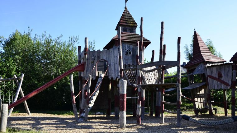 Ein Spielplatz mit einer Holzspielburg und verschiedenen Rutschen. Umgeben von grünen Bäumen und klarem Himmel.