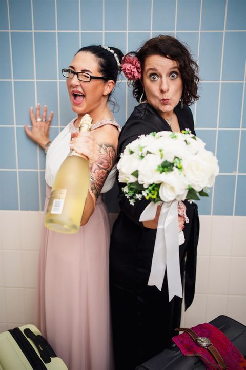 Twee vrouwen staan vrolijk samen, de een met een champagnefles en de ander met een bloemenboeket. Ze vieren duidelijk een bijzondere gelegenheid.