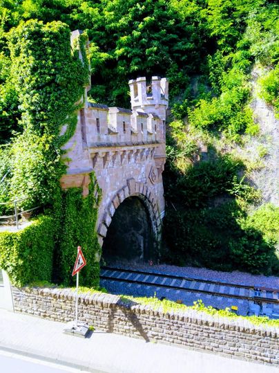 Eine alte Brücke mit Zinnen, die von grünen Pflanzen bedeckt ist. Darunter verläuft eine Eisenbahnstrecke.