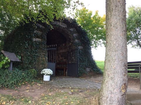 Eine kleine, mit Efeu bewachsene Kapelle steht unter Bäumen. Vor dem Eingang steht ein Blumentopf mit weißen Blumen.