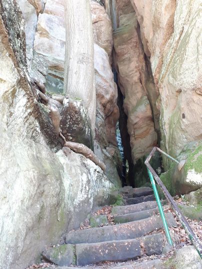 Ein schmaler Pfad zwischen hohen Felsen führt zu einer Treppe. Die Steine sind mit Moos bewachsen und die Umgebung wirkt naturnah.