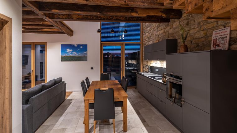 Eine moderne Küche mit einem großen Esstisch und Essbereich. Die Wände sind aus Stein und Holz, und große Fenster sorgen für viel Licht.