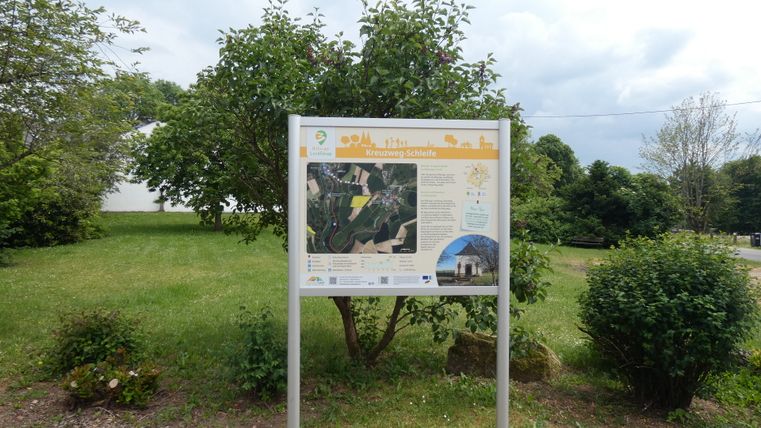 Panneau d'information sur la boucle du chemin de croix à Sefferweich, entouré d'arbres et d'arbustes.
