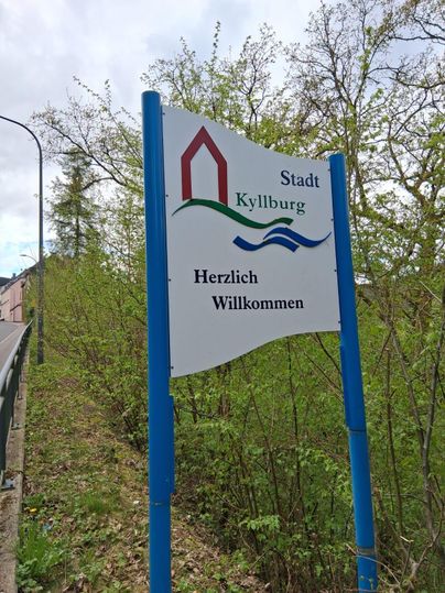 Ein Willkommen-Schild der Stadt Kyllburg. Das Schild zeigt eine grüne Landschaft mit Bäumen im Hintergrund.