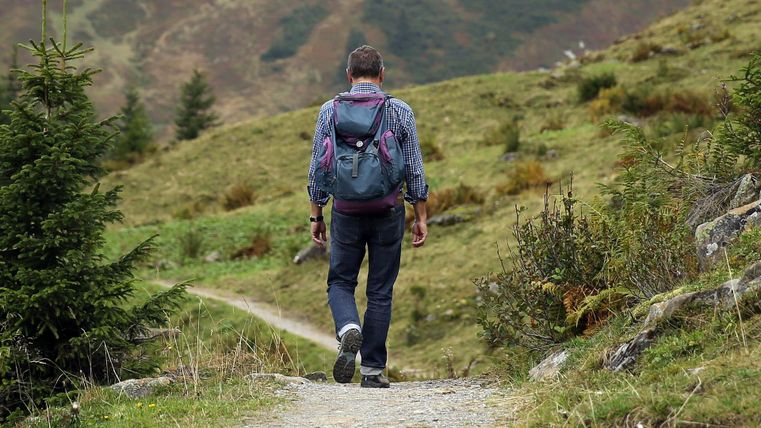 Ein Wanderer mit einem Rucksack geht auf einem schmalen Weg durch eine grüne, bergige Landschaft. Im Hintergrund sind Bäume und Hügel zu sehen.