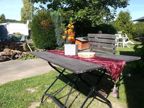 Ein einfacher Holztisch mit einer roten Tischdecke steht im Freien. Auf dem Tisch befindet sich ein Blumengefäß und im Hintergrund sind Bäume und andere Gartenmöbel zu sehen.