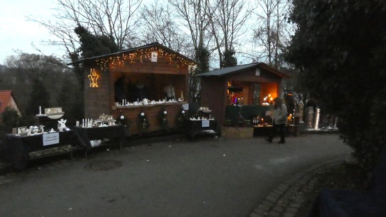 Ein Weihnachtsmarkt mit zwei geschmückten Ständen und festlicher Beleuchtung. Umgeben von Bäumen und einem schmalen Weg.