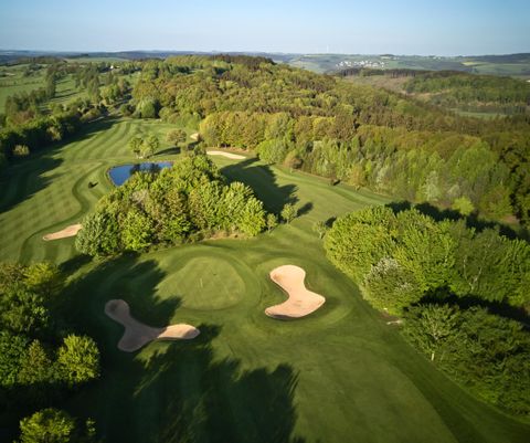 Eine weitläufige Golfanlage mit sanften Hügeln, mächtigen Bäumen und einem blauen Teich. Im Vordergrund sind mehrere Sandbunker sichtbar.