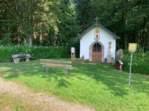 Kleine Kapelle im Wald mit Bank und Infotafel.